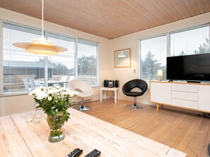 A modern living room with a wooden floor, a dining table with a vase of flowers, a TV on a cabinet, and large windows with blinds.