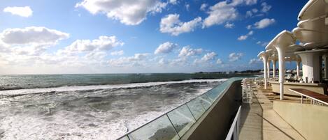 Een strandboulevard met een glazen balustrade met uitzicht op de oceaan, een rij witte parasols en een helderblauwe lucht.