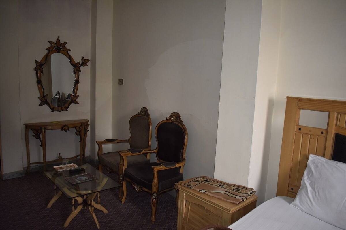 A hotel room with a wooden bed, a small bedside table, a chair, a glass-top table, and a mirror on the wall.