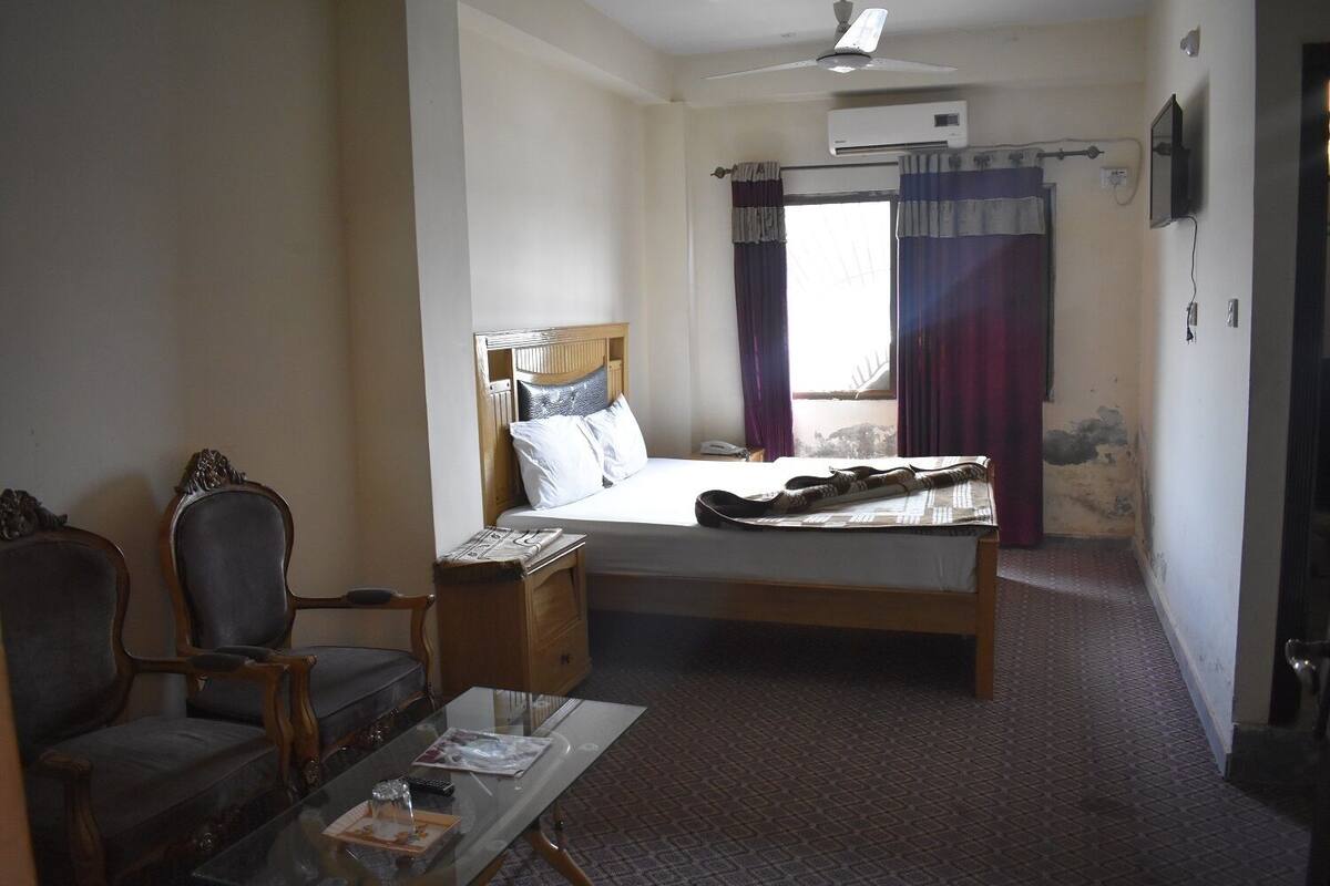 A hotel room with a bed, a wooden nightstand, a chair, a glass table with a remote control, and a window with curtains.