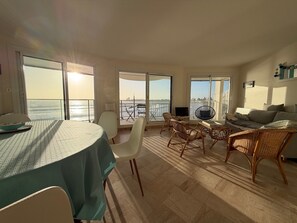 Un vaste espace de vie comprenant une table à manger, des chaises, un canapé et un téléviseur. De grandes fenêtres offrent une vue sur la mer et le paysage urbain.