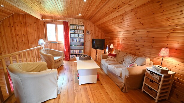Ein gemütliches Interieur einer Holzhütte mit Sofa, Sesseln, Bücherregal, Fernseher und Lampe.