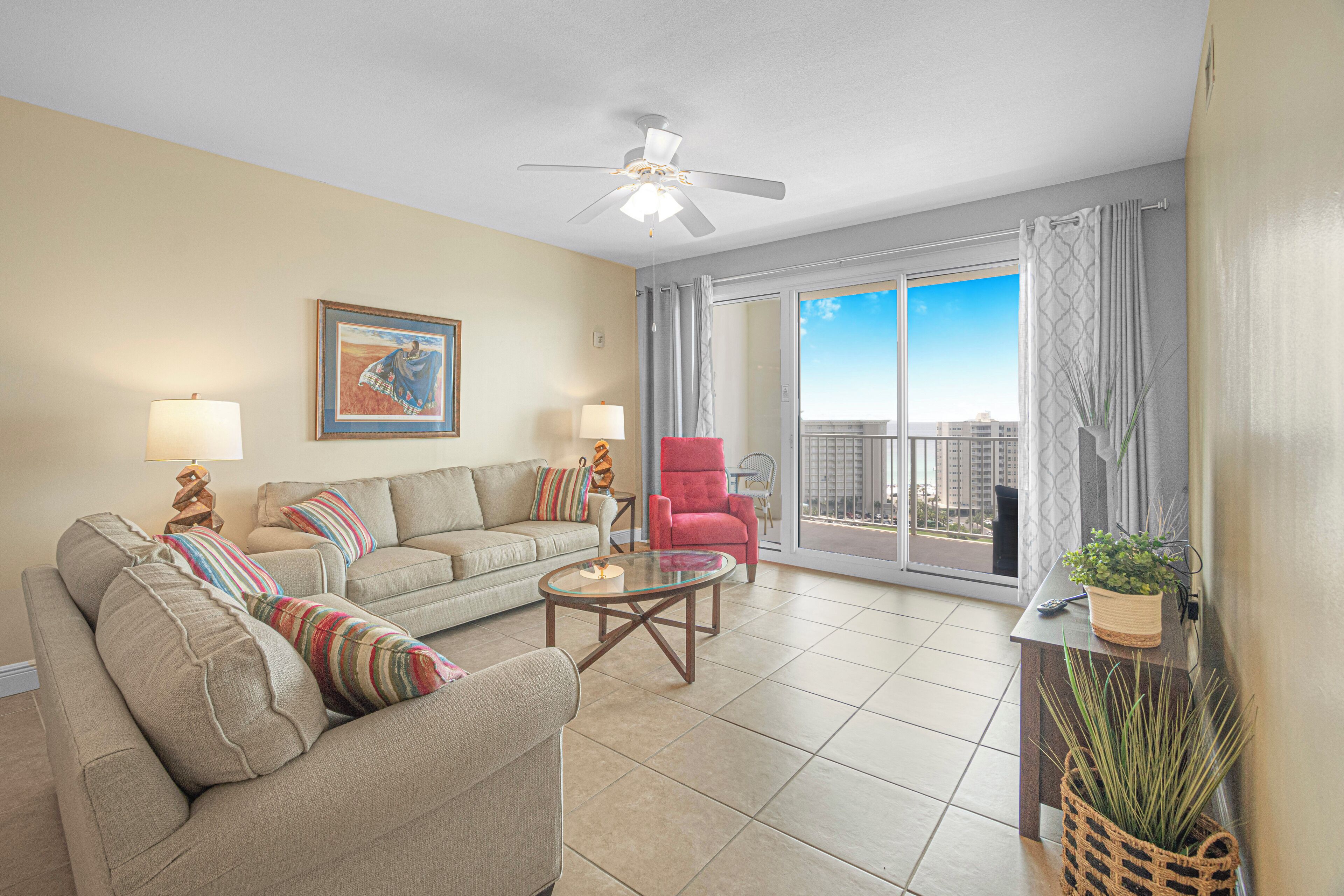 A spacious living room with a sofa, armchair, and a glass coffee table. There is a ceiling fan, a painting on the wall, and a view of the city through large windows.