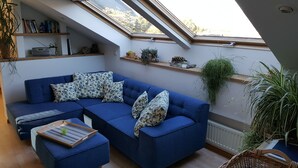 A modern living room with a blue sectional sofa, a small wooden table, and a large window with a view of trees. - Above the rooftops of Lana - Blue apartment with terrace and sauna - viaDolceVita (Lana)
