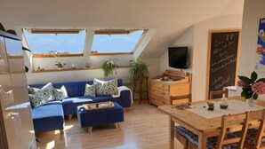 A cozy living room with a blue sofa, wooden dining table, and a TV. There are skylights and a chalkboard with writing. - Above the rooftops of Lana - Blue apartment with terrace and sauna - viaDolceVita (Lana)