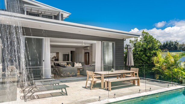 A modern house with a swimming pool, a dining area with outdoor furniture, and a large glass door leading to a living room.