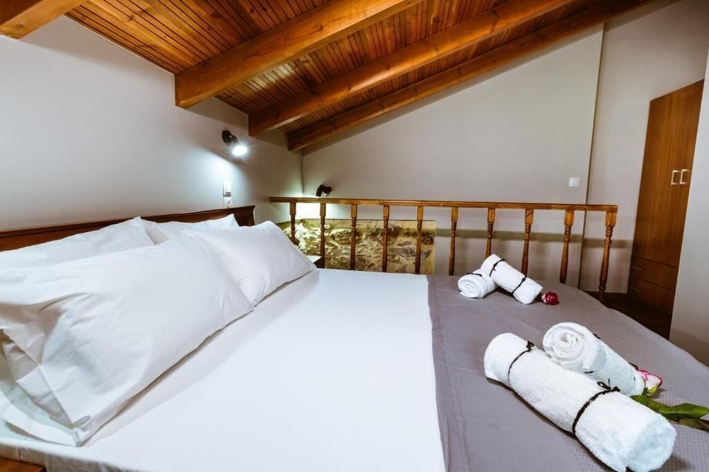 Un dormitorio con techo de madera, una cama con ropa de cama blanca, un balcón con barandilla de madera y una pared de piedra.