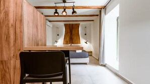 A modern, minimalist room with a wooden dining table, a black chair, a bed with a white headboard, and a large window providing natural light. - Ellafos Traditional Living (Kissamos)