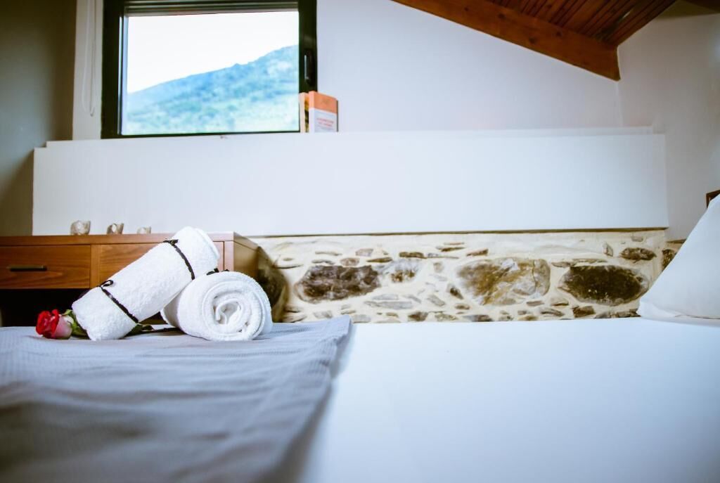 Hotellihuone, jossa on kiviseinä, puukatto, valkoisilla lakanoilla varustettu sänky, rullalle taitettu pyyhe, punainen ruusu ja ikkuna, josta avautuu näkymä vuorille.