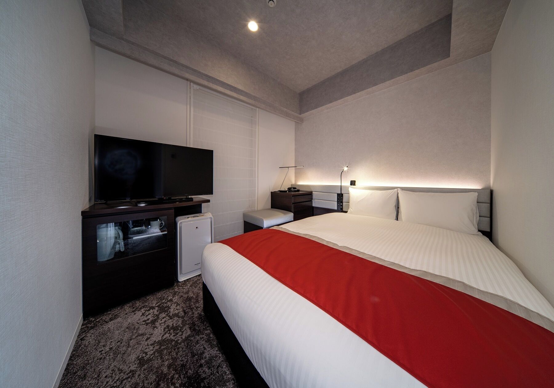 A hotel room with a bed, a television, a desk with a lamp, and a small refrigerator.