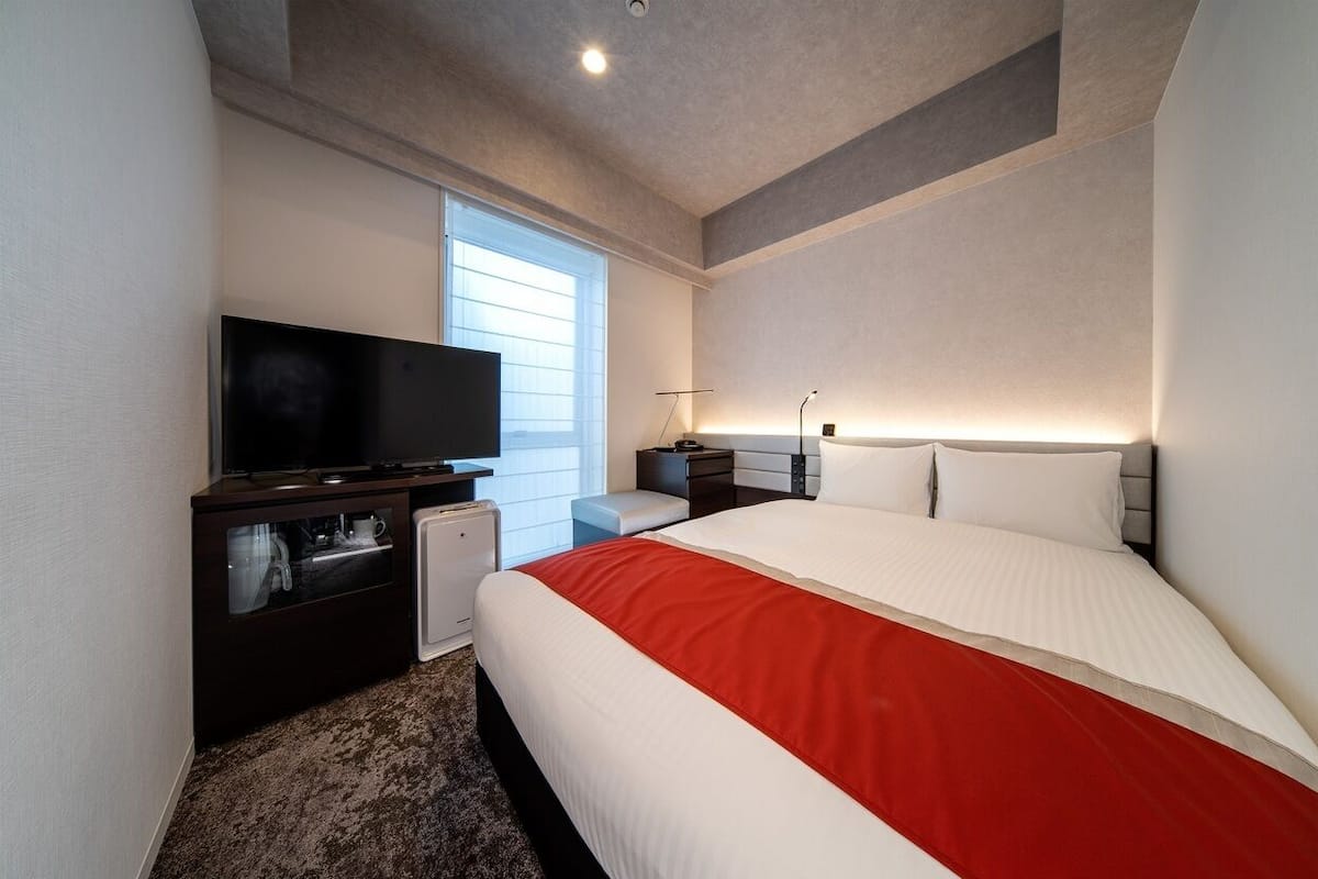A modern hotel room with a large bed, a flat-screen TV, a small desk, and a window with blinds.