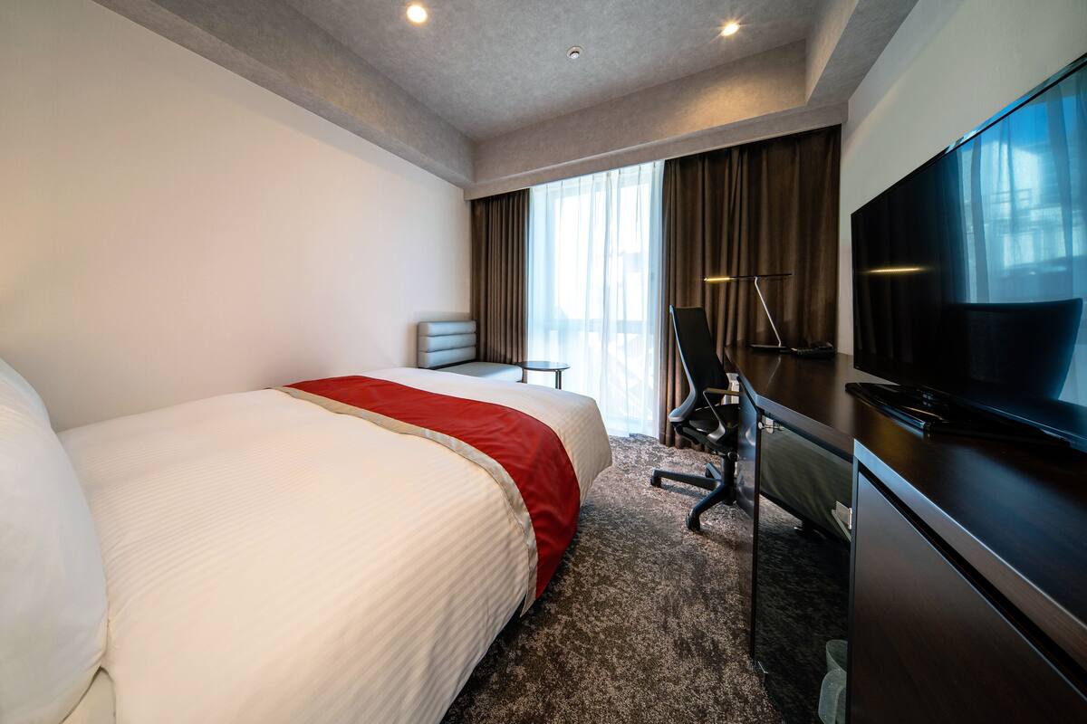 Een hotelkamer met een groot bed, een bureau met stoel, een televisie en een raam met gordijnen.