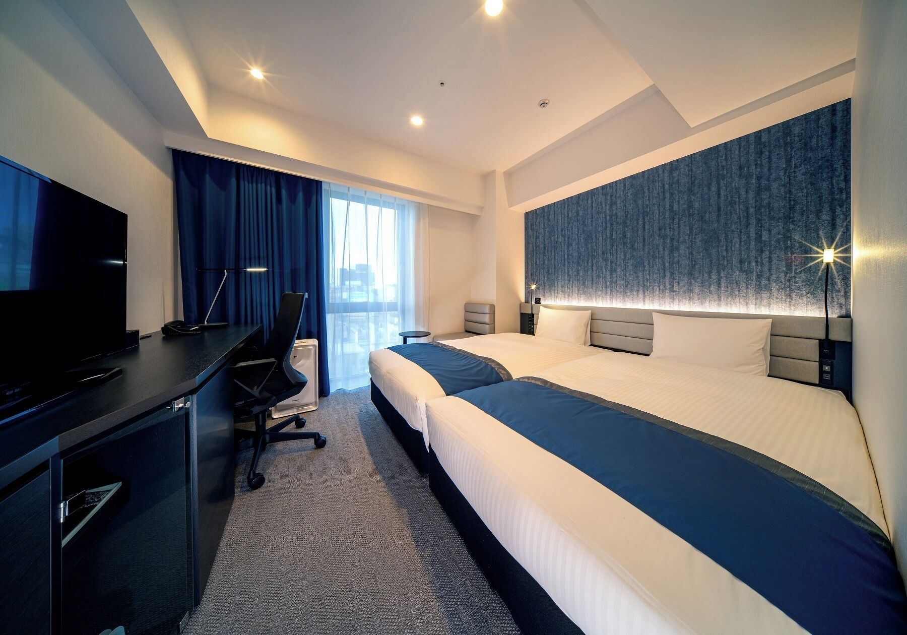 A modern hotel room with a large bed, a desk with a flat-screen TV, and a chair.