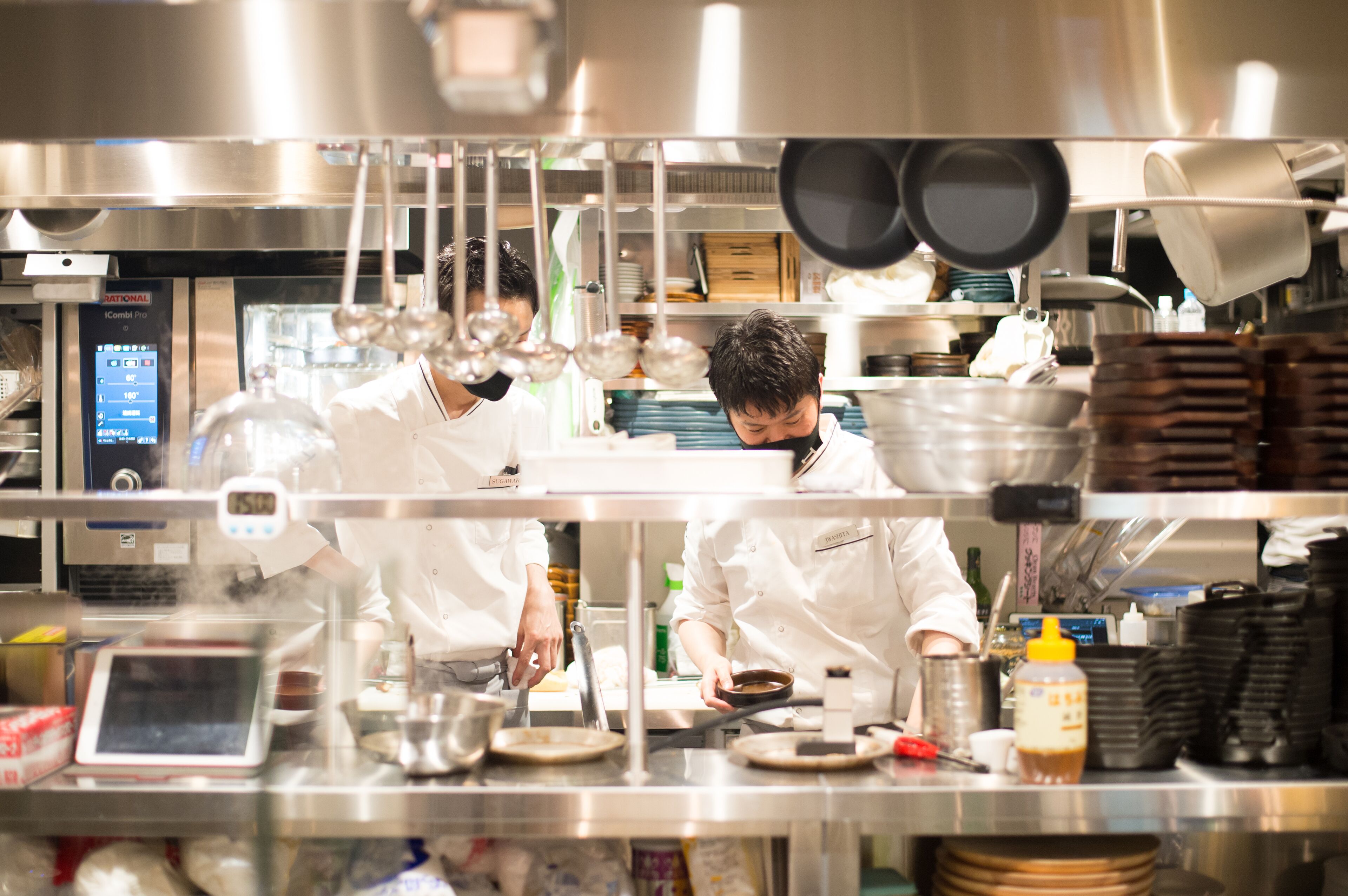 A professional kitchen with stainless steel surfaces, hanging pots, and a digital display.