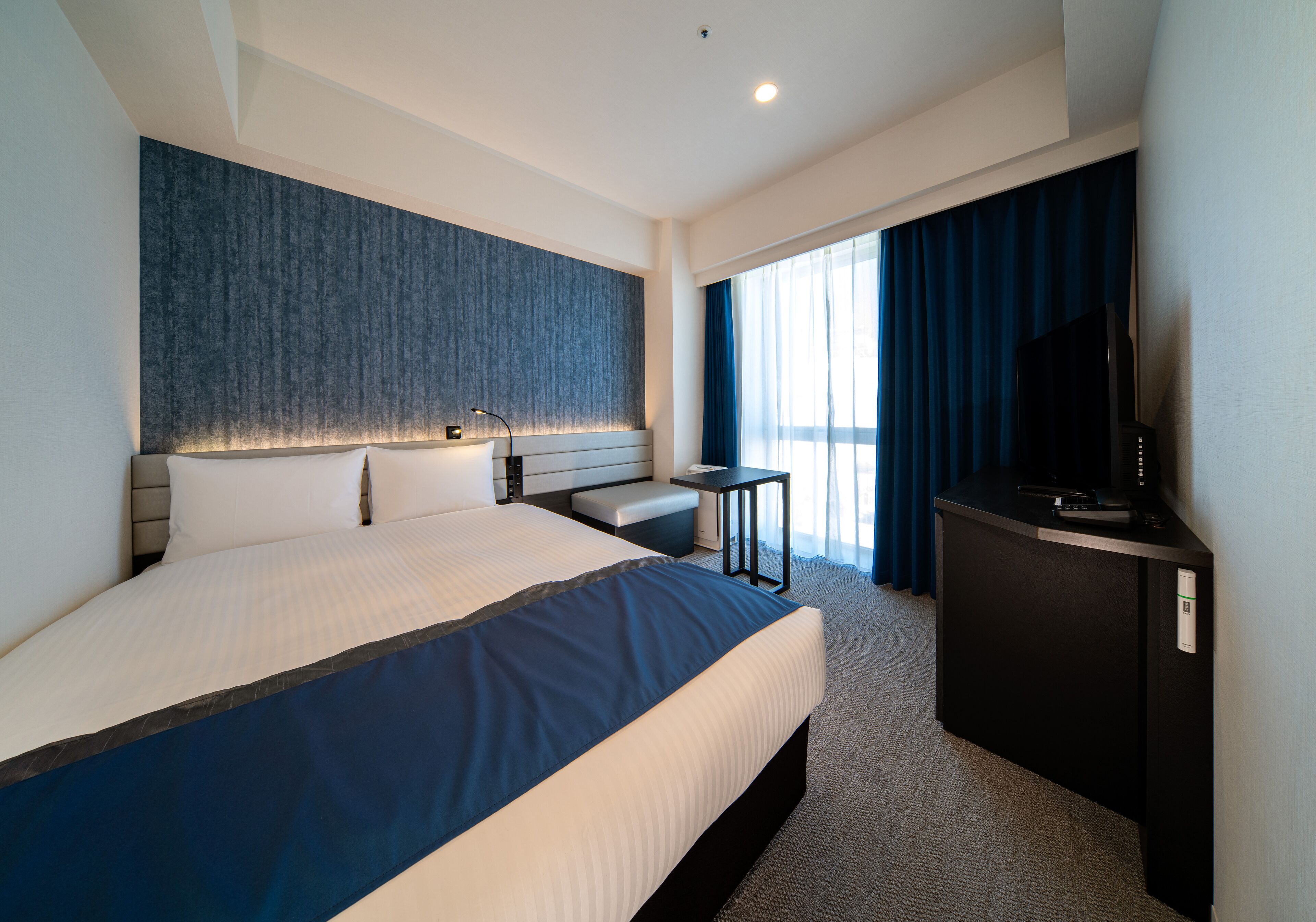 A modern hotel room with a large bed, a desk, a television, and a view of the outdoors through large windows.