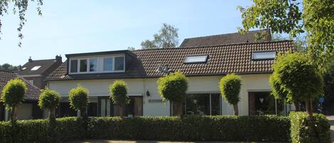 A building with a tiled roof, a sign reading 'DE WINGERD,' and a paved courtyard with hedges and trees.