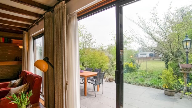 A cozy room with a wooden ceiling, a red sofa, a wooden table with a vase of tulips, and a view of a garden through a glass door.