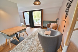 A modern living room with a wooden dining table, grey armchairs, a sofa, and a small table. There is a large window with a view of outdoor furniture. - Apartment in Vielsalm Near Les Doyards Park (Vielsalm)