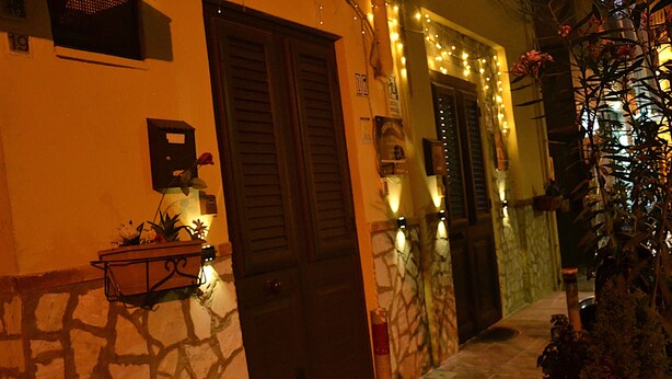 A building with stone walls and wooden doors, a small outdoor seating area with a table and chairs, and string lights along the entrance. - casa Del Vicolo (Palermo)