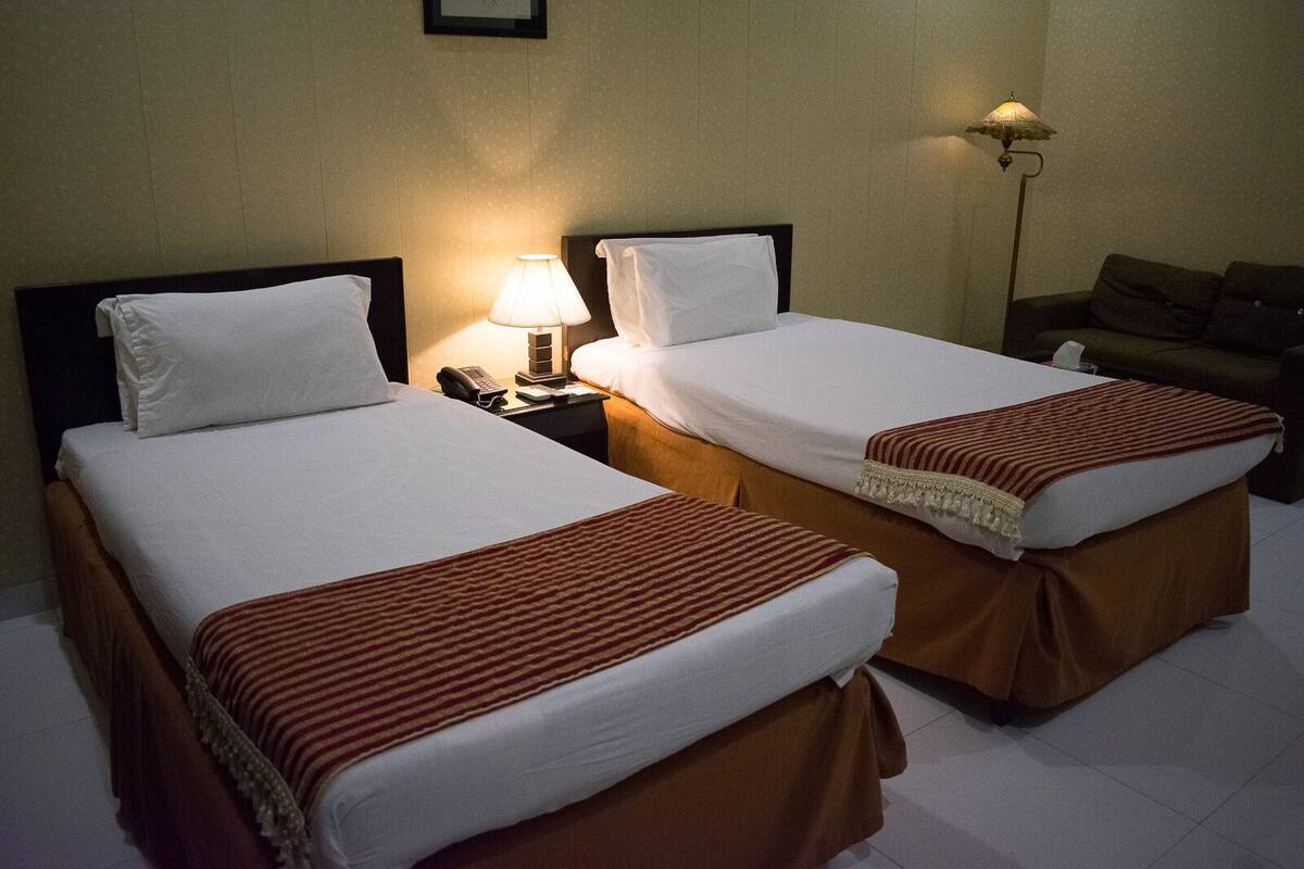 Dos camas individuales con sábanas blancas y cabeceros marrones, una lámpara de noche y un sofá en una habitación de hotel.