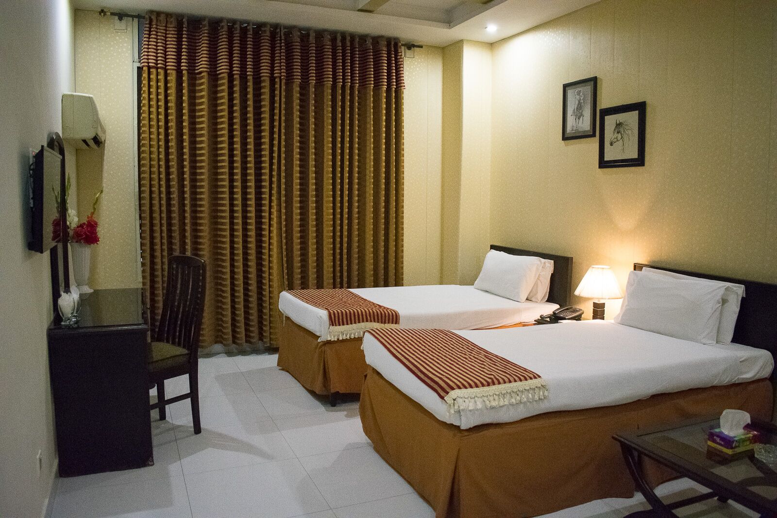 Habitación de hotel con dos camas, un escritorio, una silla, una lámpara y dos cuadros en la pared.