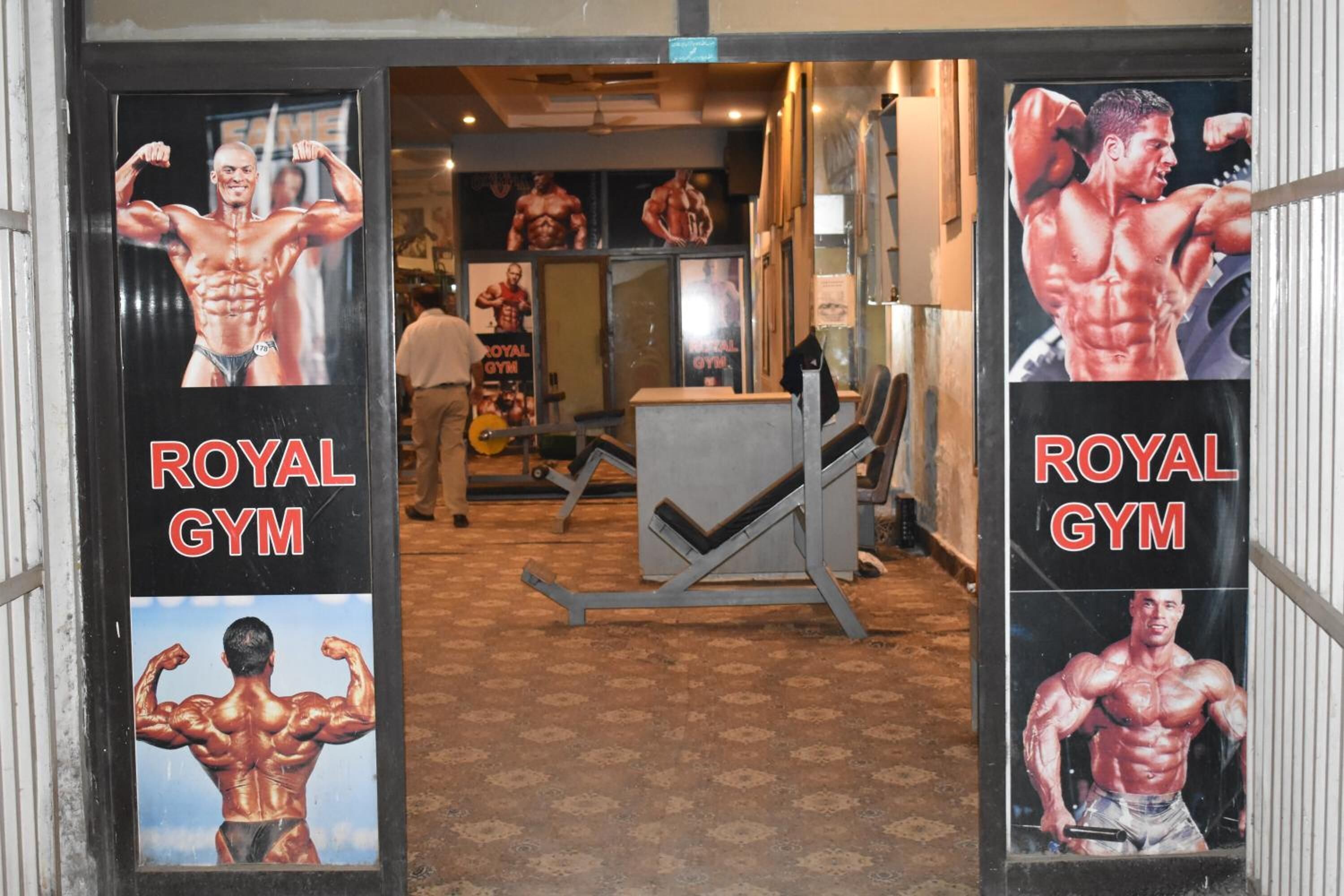 Royal Gym -kuntosalin sisäänkäynti, jossa on seinillä kehonrakennusaiheisia julisteita ja keskellä painonnostopenkki.
