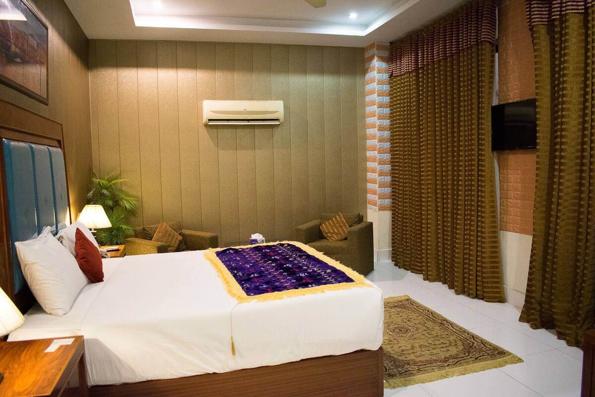 Habitación de hotel con cama, mesita de noche, lámpara, sillón, pequeña mesa, planta, equipo de aire acondicionado, televisión y cortinas.