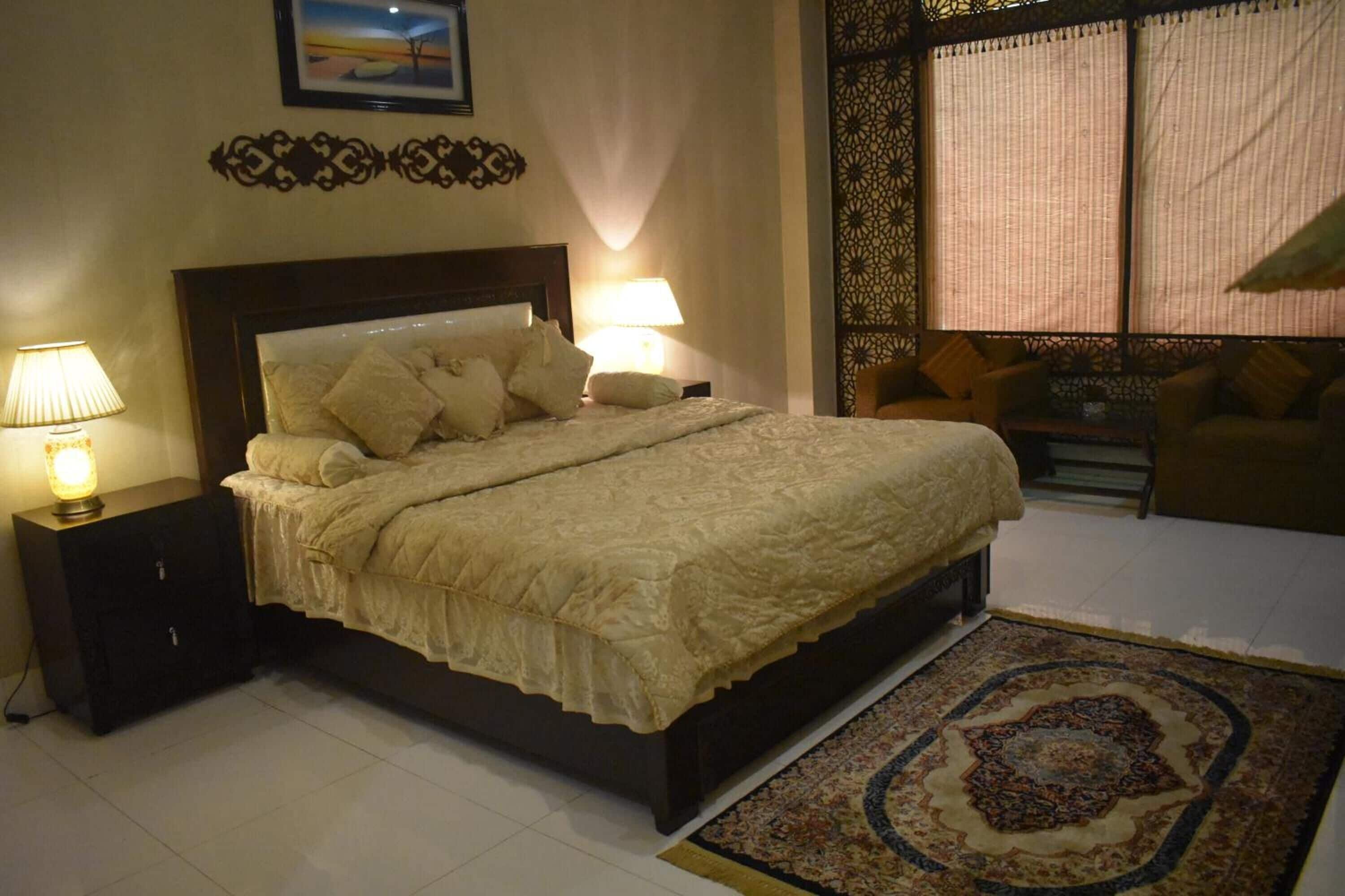 Un dormitorio ordenado con una cama grande, dos mesitas de noche con lámparas, un sofá y un ventanal decorativo con cortinas.