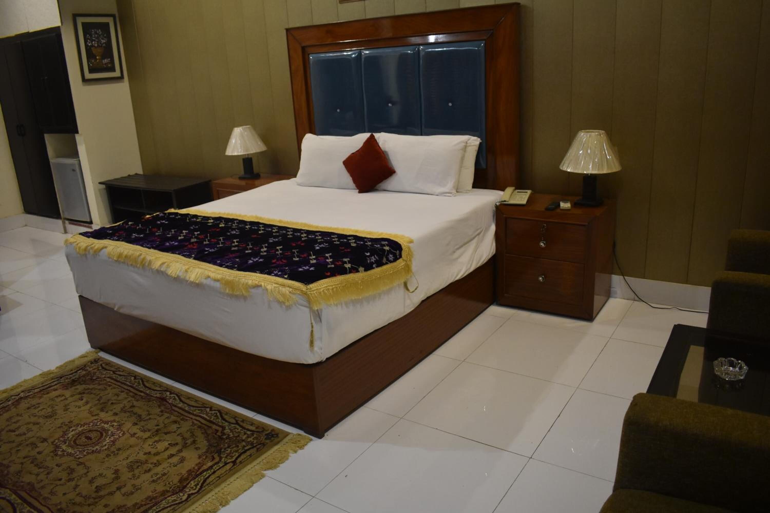 A neatly made bed with a white comforter and a decorative throw, a wooden nightstand with a lamp, and a small table with a glass object.