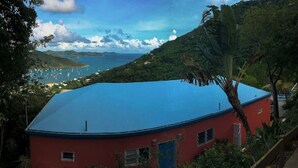 A red building with a blue roof, surrounded by lush greenery and a view of the sea with boats. - "Someday Soon" Has Arrived! (St John)