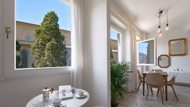Ein Zimmer mit Essbereich, einem Fenster mit Blick auf ein Gebäude und einen Baum sowie einer Zimmerpflanze.