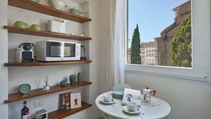 Ein Zimmer mit Holzböden, die Küchengeräte, eine Uhr und eine Flasche beherbergen. Ein Tisch mit Tassen und Untertassen, und ein Fenster mit Blick auf Gebäude und Bäume.