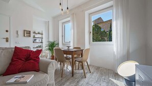 Ein modernes Wohnzimmer mit einer Couch, einem Esstisch mit Stühlen, einer TV-Konsole und einem Fenster mit Blick auf ein Gebäude und Bäume.
