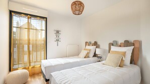 Une chambre moderne avec deux lits, une grande fenêtre ornée de rideaux légers, un luminaire en fibres tressées et un petit pouf rond.