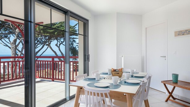 Un coin repas avec une table dressée pour quatre personnes, une petite table d’appoint en bois et une vue sur la mer, visible à travers de grandes portes vitrées.