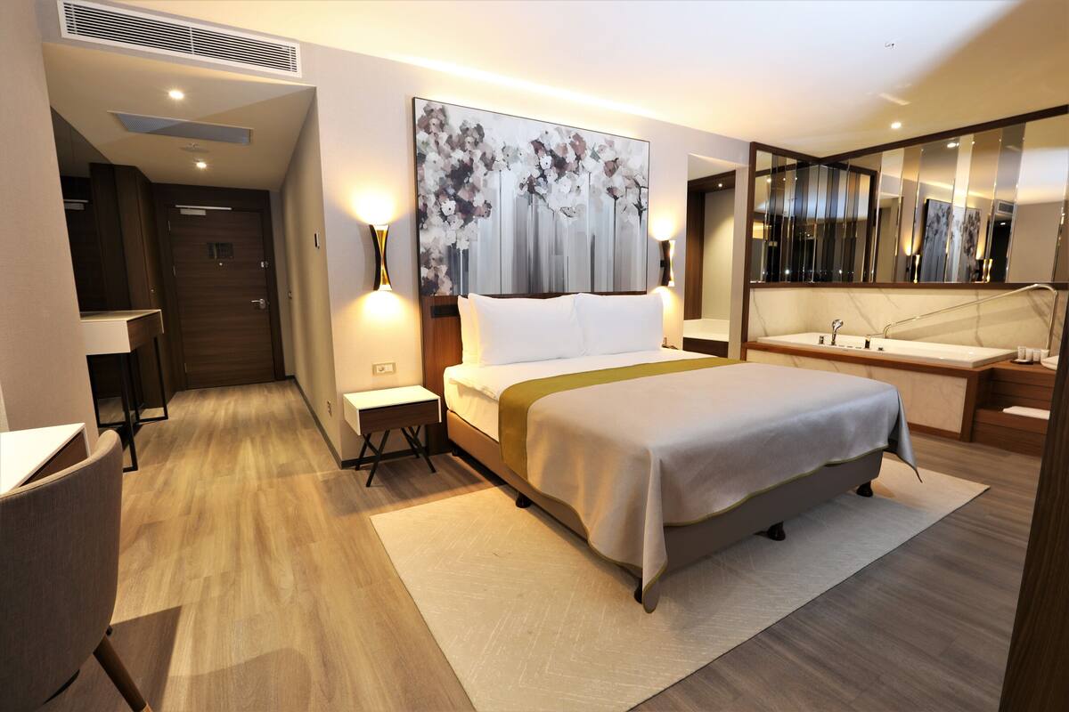 A modern hotel room with a large bed, bedside tables, a desk, a chair, a bathroom with a sink and mirror, and a large painting on the wall.