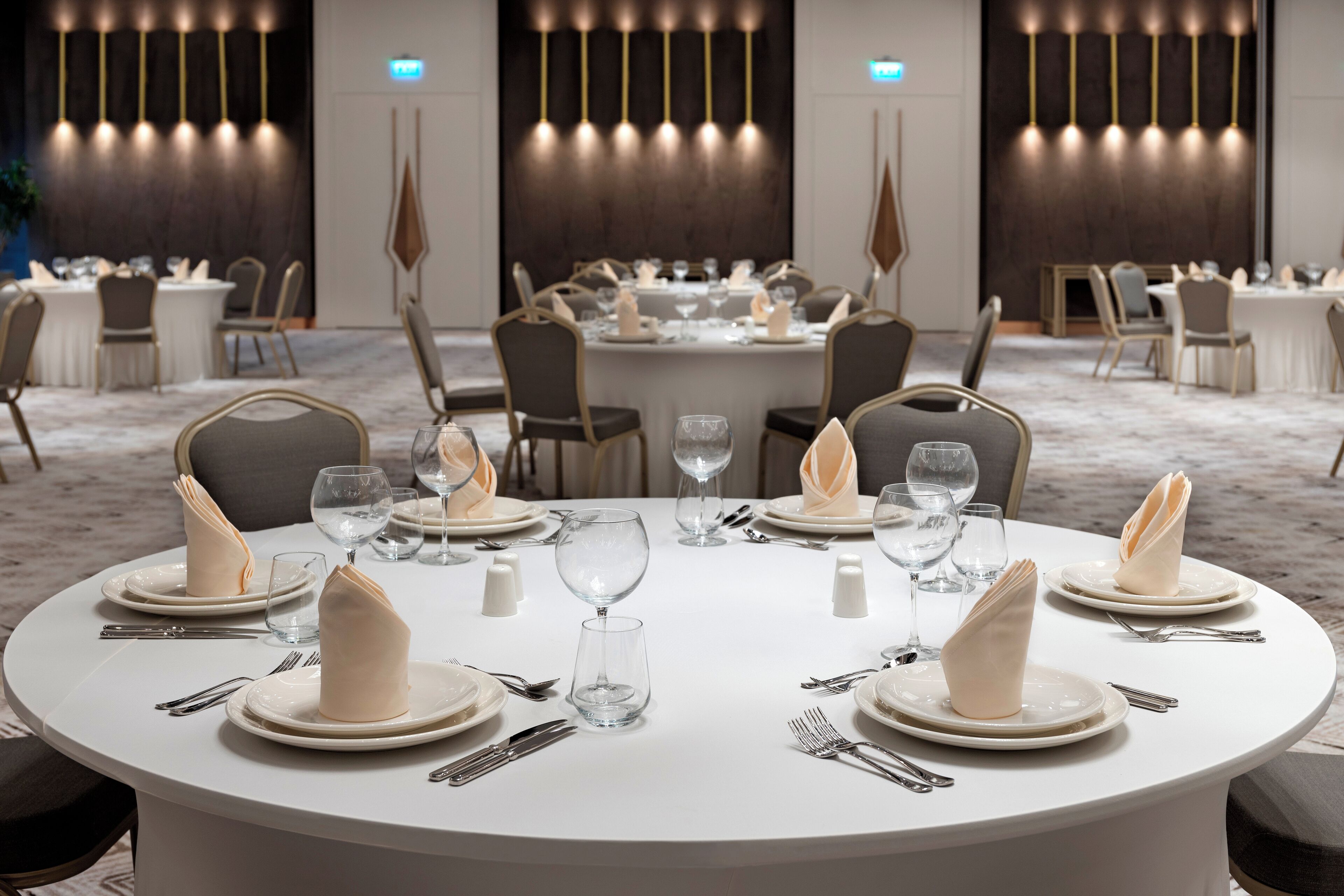 Une table de salle à manger ronde, dressée avec de l’argenterie blanche, des serviettes pliées et des verres à vin, entourée de chaises dans un espace de restauration moderne d’un hôtel, avec un sol en marbre et des luminaires suspendus.