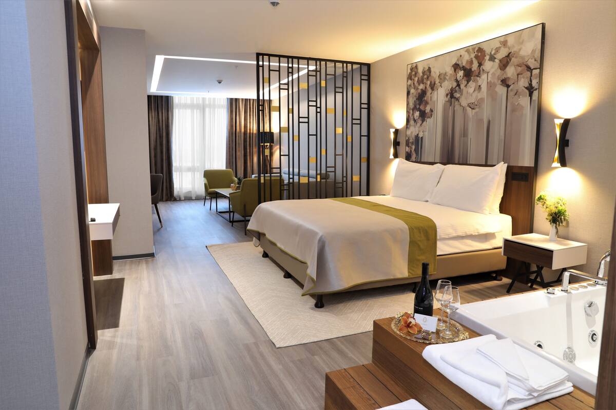 A modern hotel room with a large bed, a sitting area with a chair and table, a bathroom with a bathtub, and a decorative wall piece.