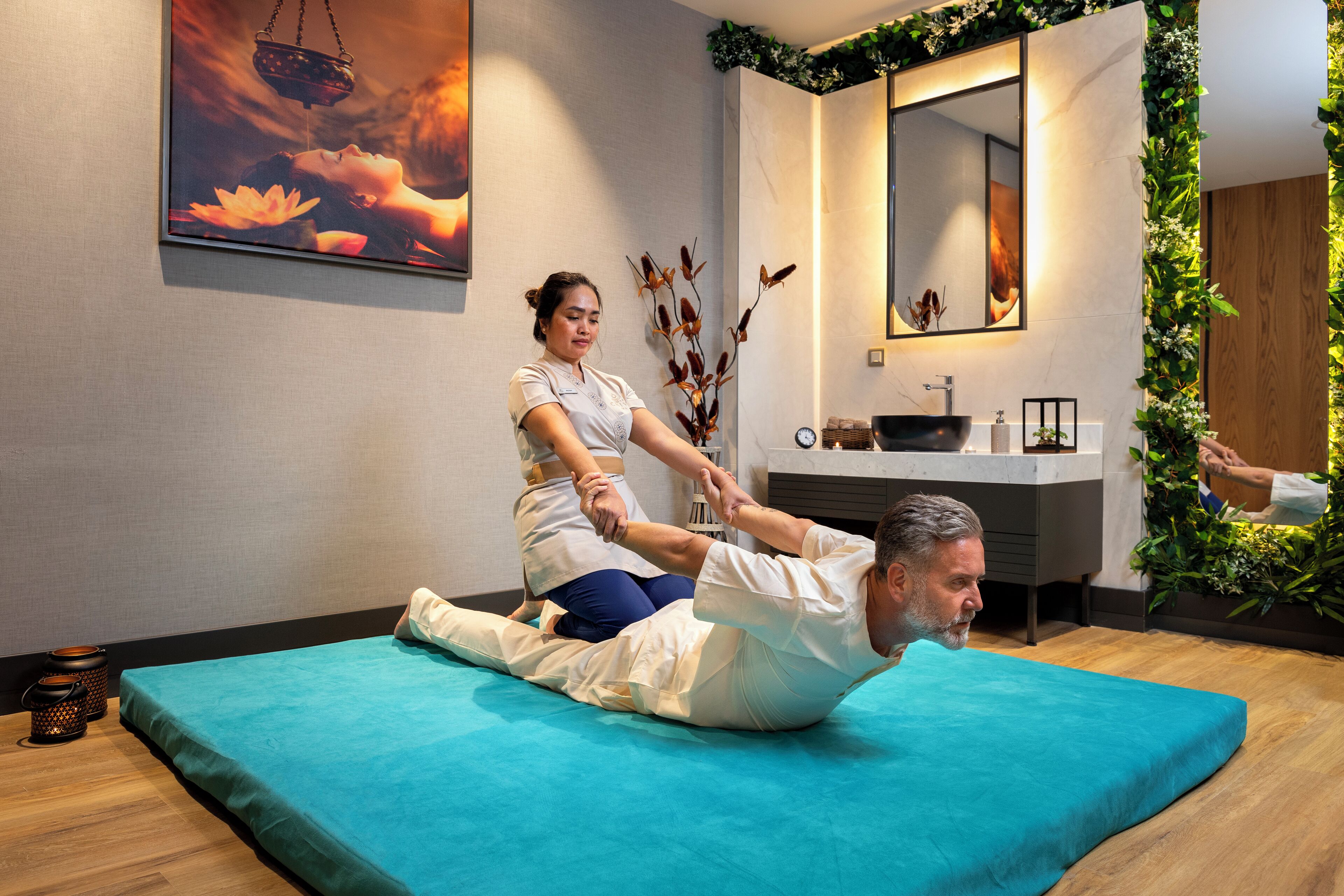 A massage therapy session in a modern room with a wooden floor, a teal massage table, a large mirror, and a wall-mounted plant.
