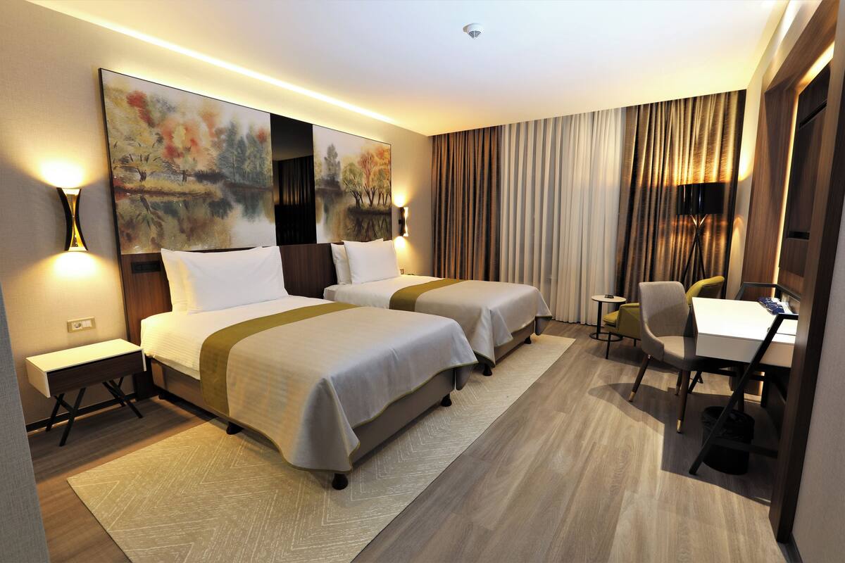 A hotel room with two beds, a desk, and a chair. There is a painting on the wall and a lamp on the nightstand.