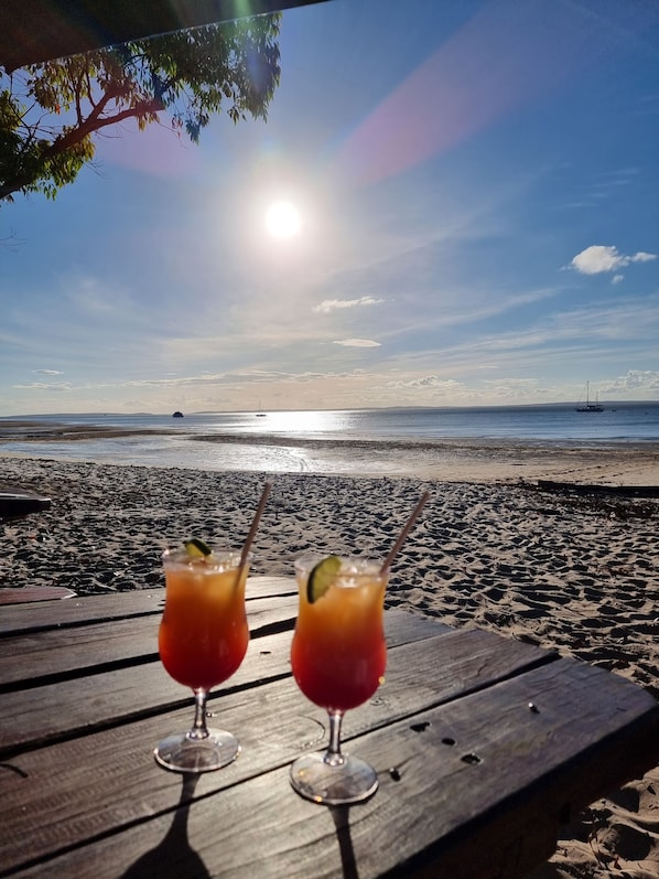 Deux boissons sur une table en bois, avec une plage et l’océan en arrière-plan.