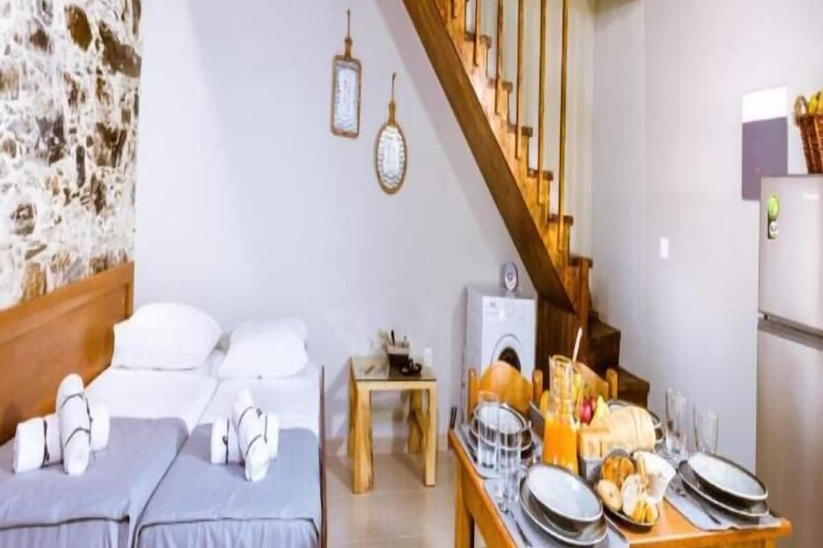 Une pièce chaleureuse avec un mur en pierre, des poutres en bois au plafond, un lit avec une literie grise, une petite table avec des éléments pour le petit-déjeuner et un escalier.