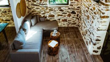 A cozy room with a stone wall, a wooden floor, a grey sofa, and two wooden stools with a book and a teapot on them.