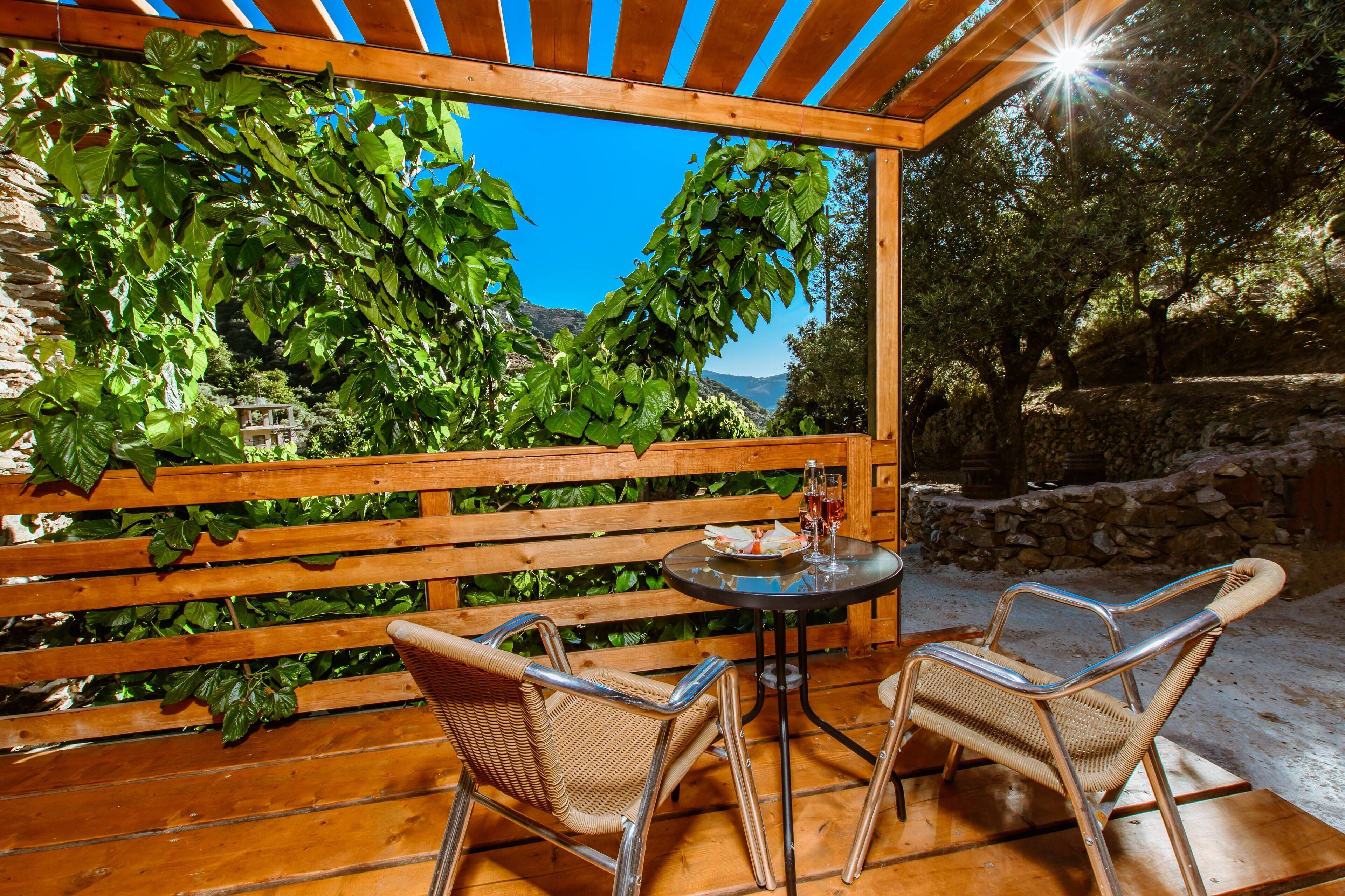 A wooden pergola with a table and two wicker chairs, overlooking a mountainous landscape.