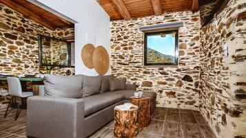 Una habitación acogedora con pared de piedra, techo de madera, un sofá, dos taburetes de madera y una ventana con vistas al exterior.