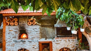 A stone-built outdoor fireplace with logs, a small wooden structure, and hanging plants. - Traditional Apartments Elafos (Kissamos)