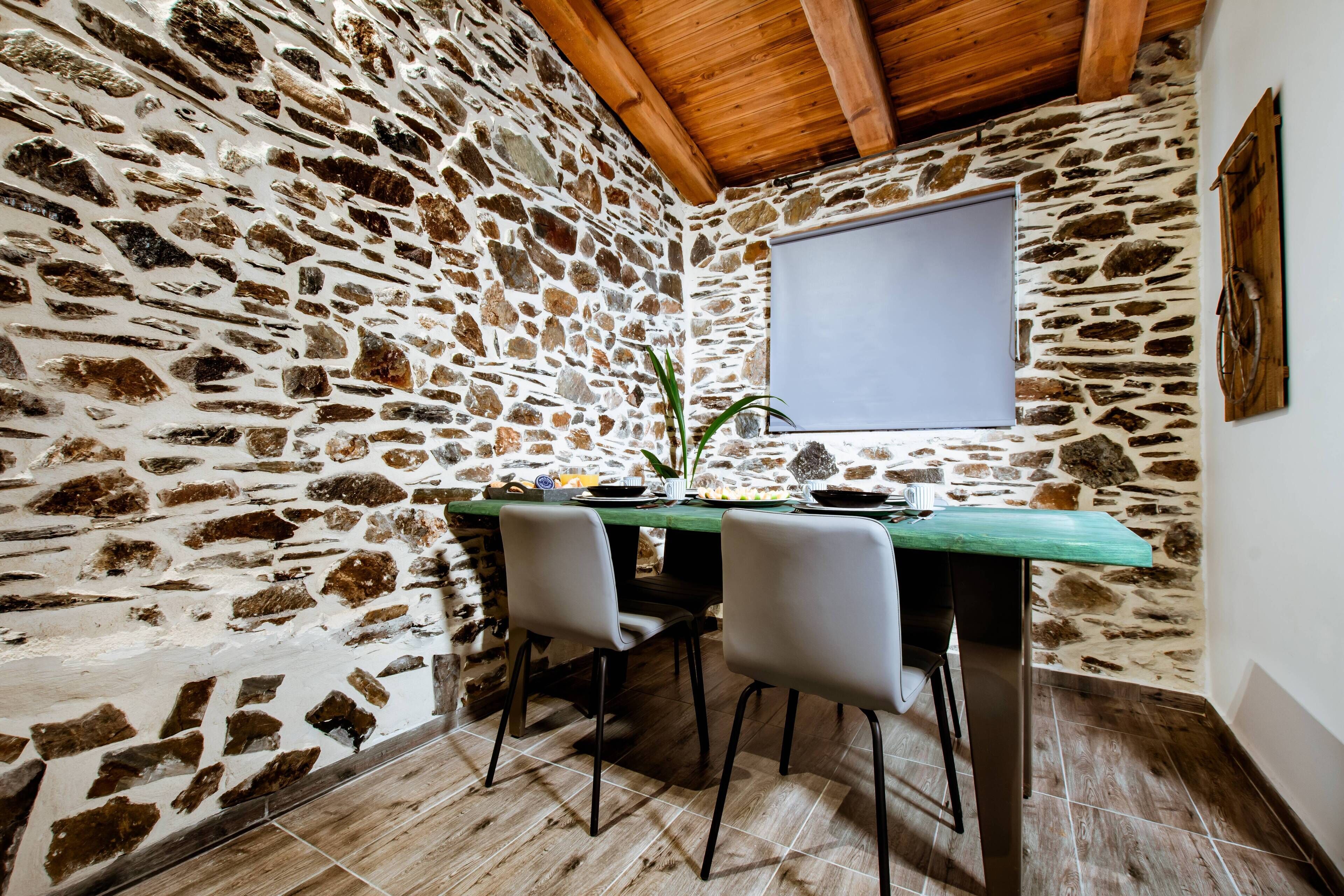 Een kamer met stenen muren, een houten plafond, een groene tafel met stoelen en een raam met een witte zonnescherm.