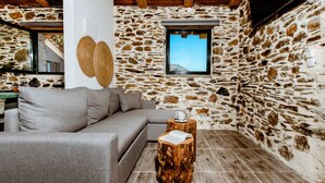 A cozy room with a stone wall, a wooden ceiling, a grey sofa, and a small wooden table with a book. - Traditional Apartments Elafos (Kissamos)