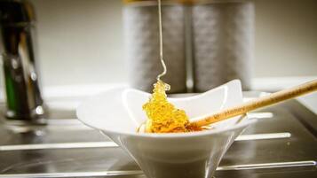 Un tazón blanco con panal sobre una cuchara, con una botella de miel y recipientes al fondo.