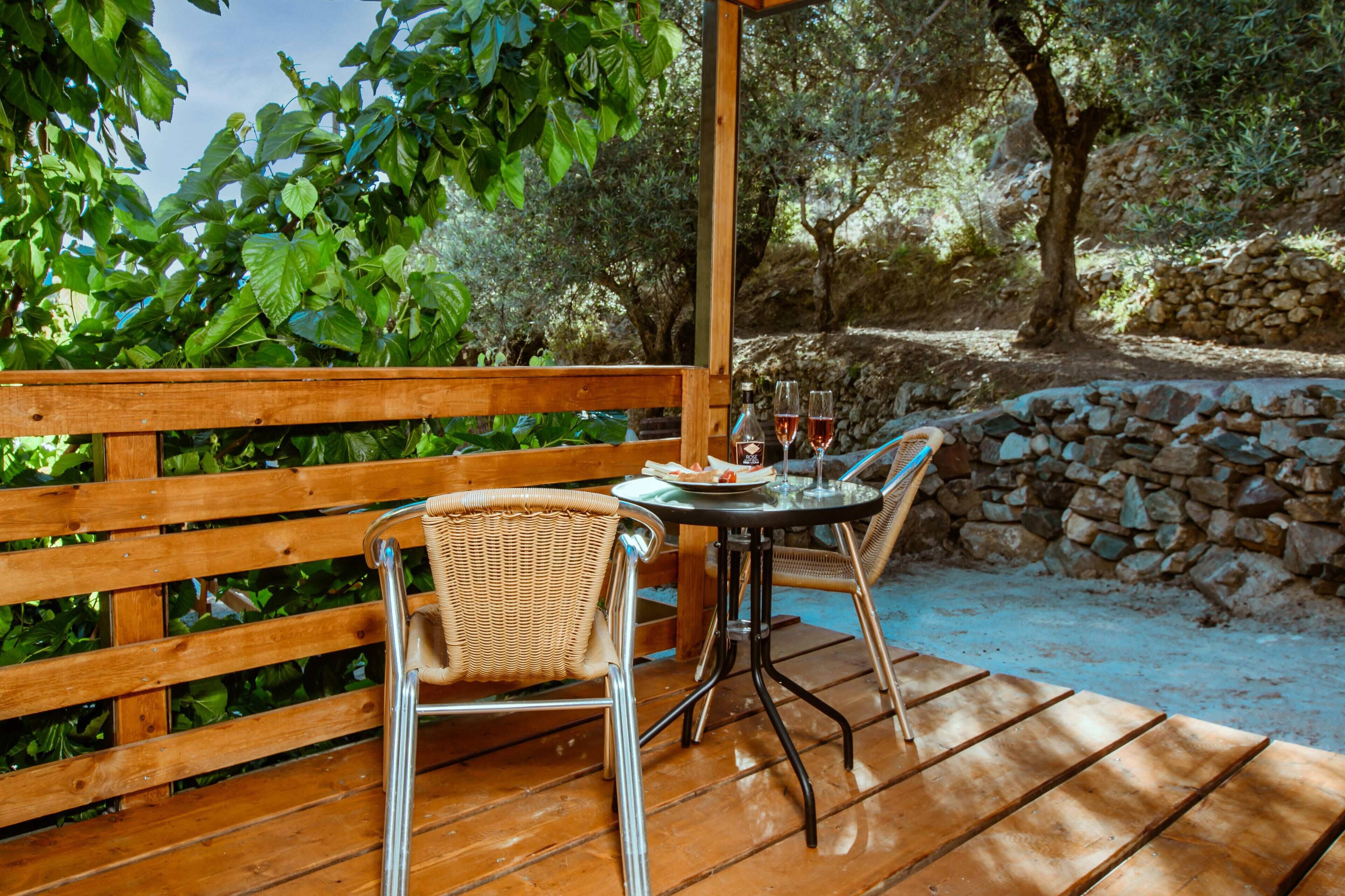 A wooden deck with a table and chairs, surrounded by greenery and stone walls.